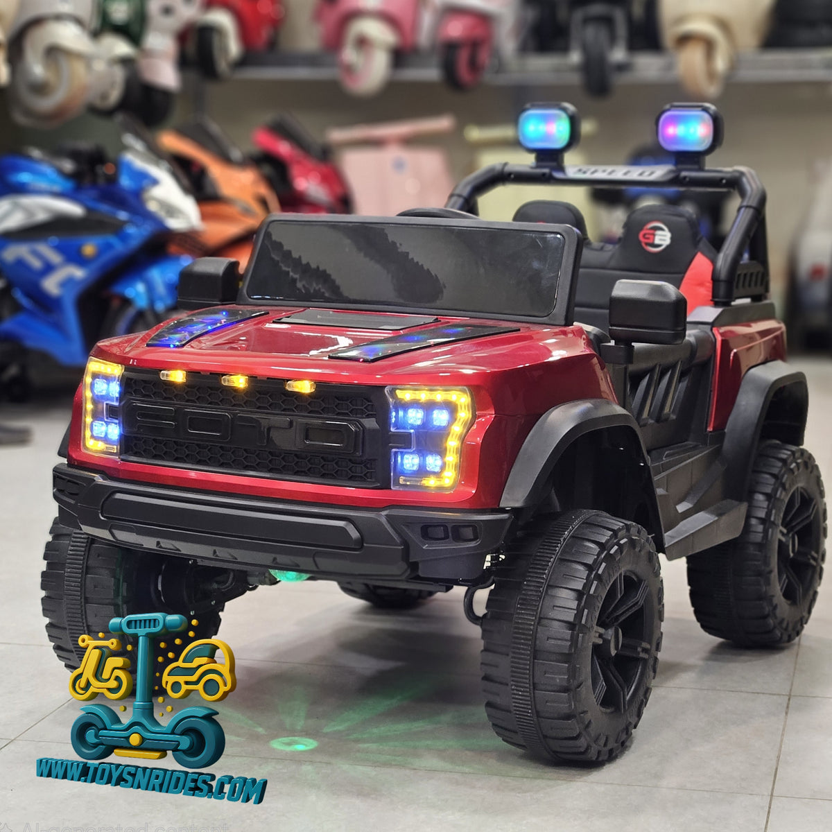 A red and black toy police car ride-on truck with flashing blue LED lights, black off-road tires, and a black roll bar, shown in a showroom setting.
