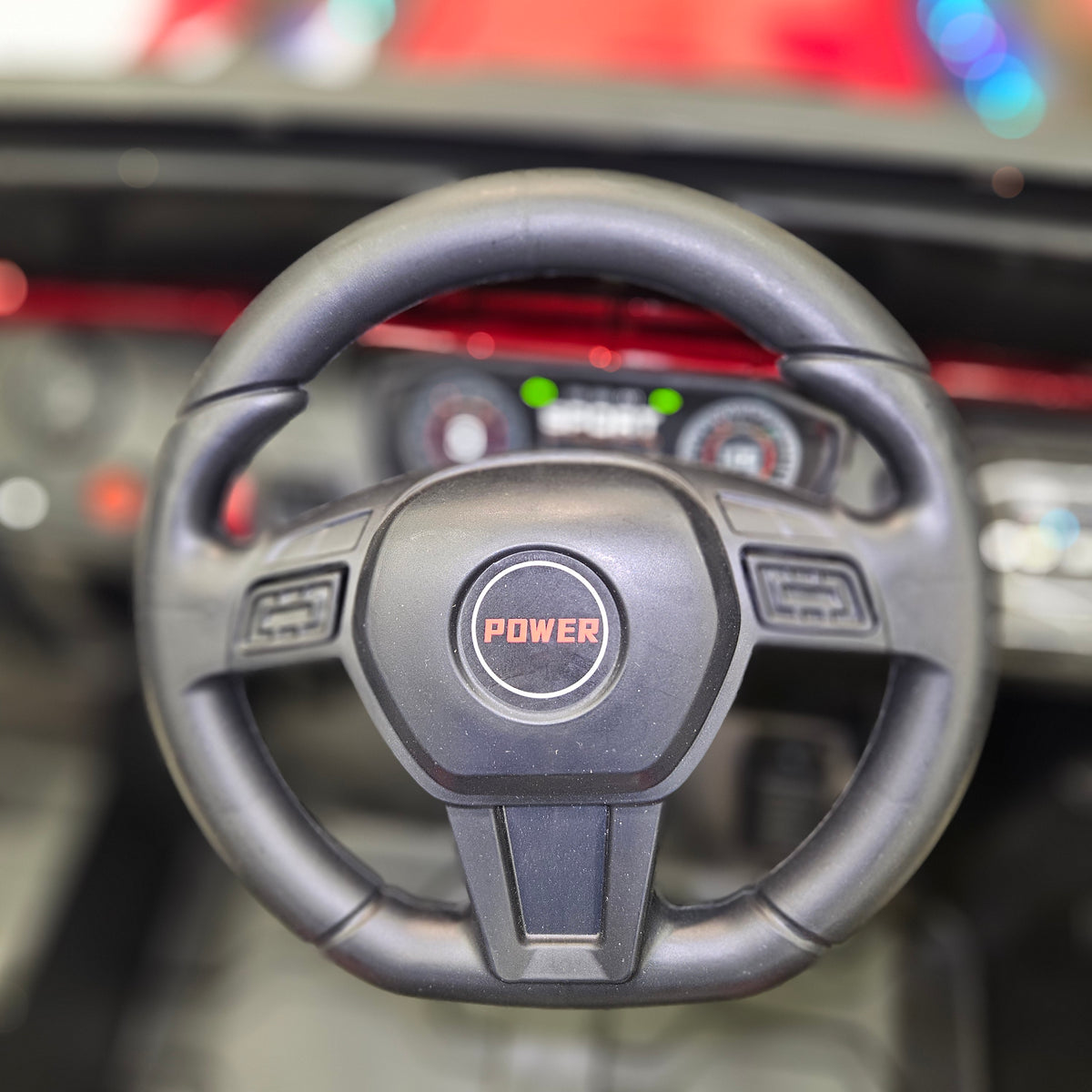 A close-up of a black steering wheel with a central logo badge that reads 'POWER' in orange, mounted inside a car dashboard with blurred instrument gauges and red ambient lighting in the background.