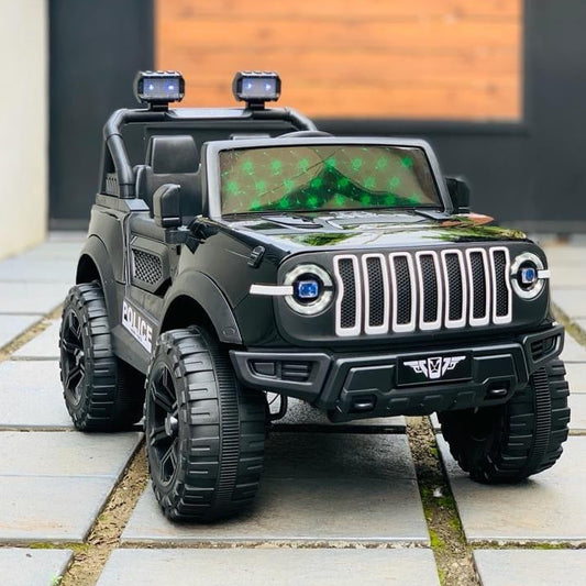 A black battery-operated remote control jeep for kids with green LED lights, positioned on a stone pathway with a wooden fence and shrubbery in the background.