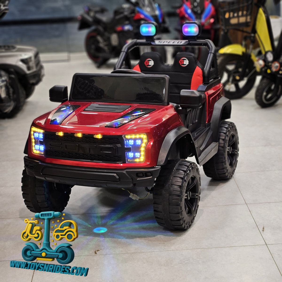 A red and black kids' ride-on toy truck with large off-road tires, LED light bars, and dual light bars on the roof, displayed in a showroom with other vehicles blurred in the background.