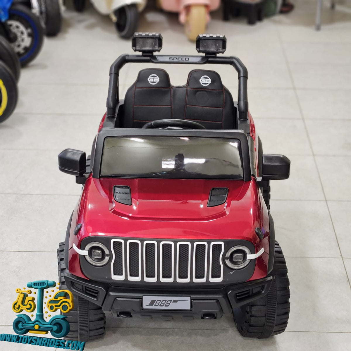 A red and black toy Jeep-style ride-on vehicle with two seats, shown from the front in a showroom setting.