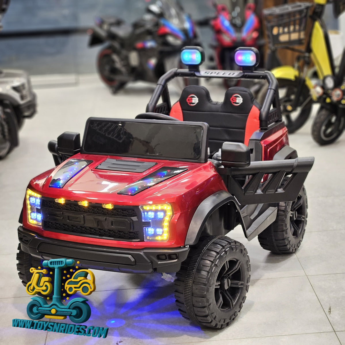 A red and black toy off-road truck with LED lights, shown in a showroom with motorcycles in the background.