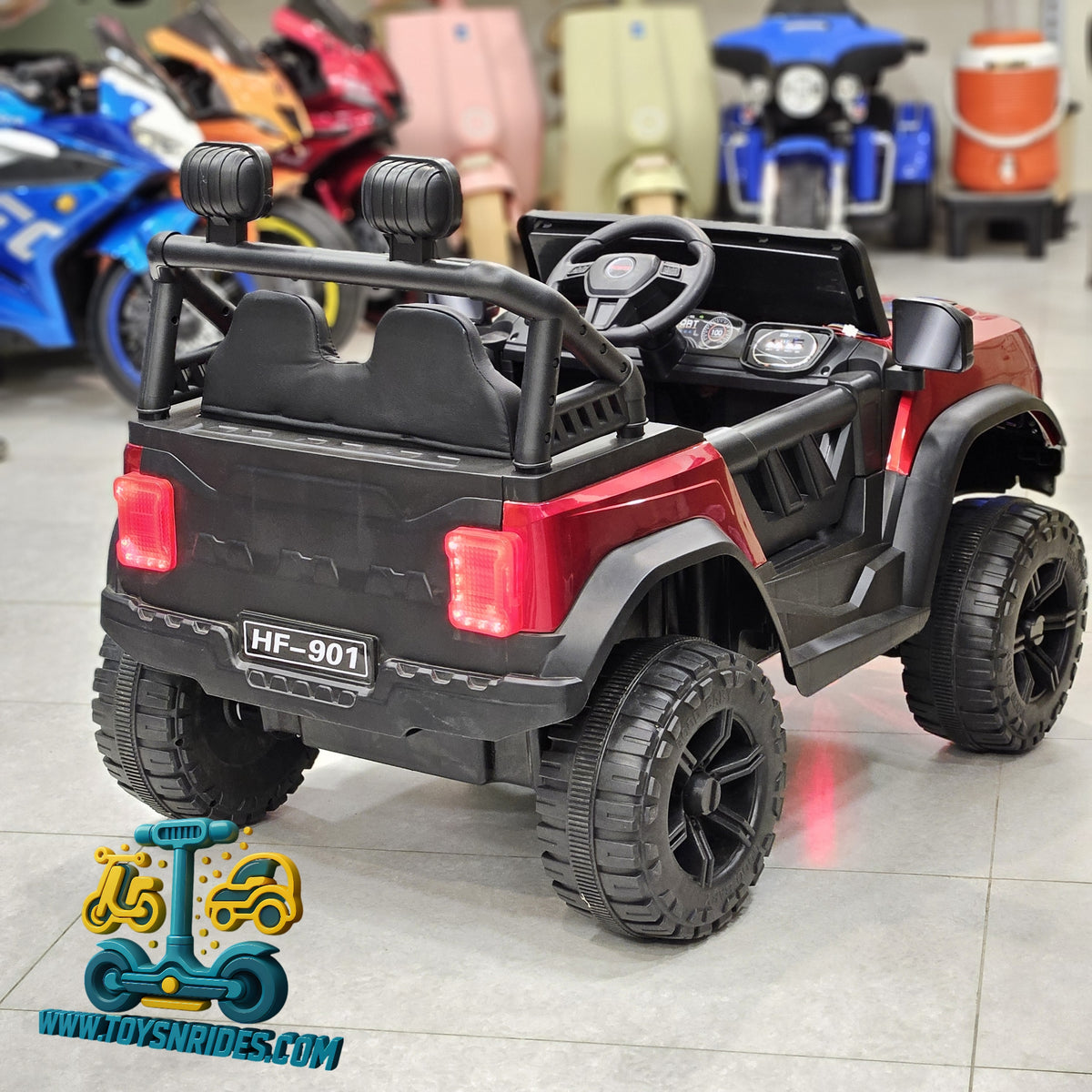 A red and black kids' electric ride-on toy jeep with large off-road tires, shown from the rear three-quarter angle in a showroom. The toy features a black roll cage, seats, and wheels, with red body panels and illuminated red taillights.