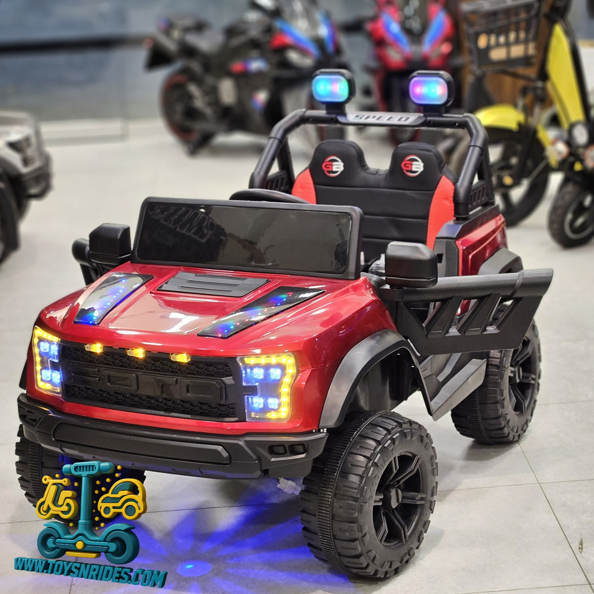 A red and black kids' ride-on toy truck with illuminated LED lights, displayed on a showroom floor with motorcycles in the blurred background.