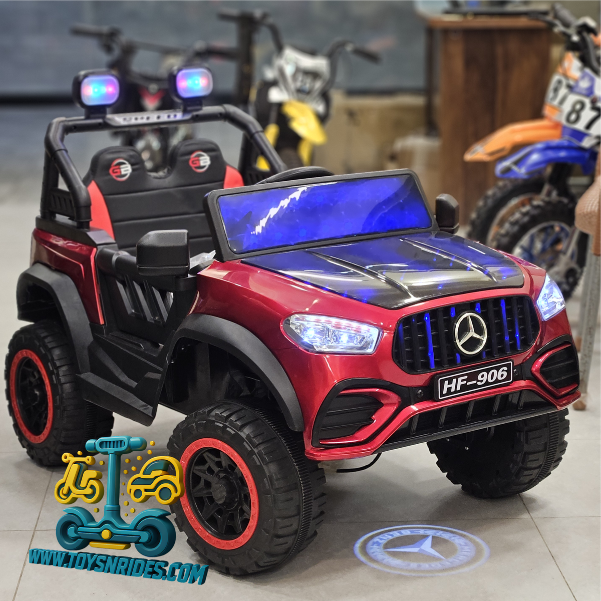 A red and black Mercedes-Benz-style children's ride-on toy truck with blue LED light bar, headlights, and a blue-tinted windshield, shown in a showroom with dirt bikes in the background.