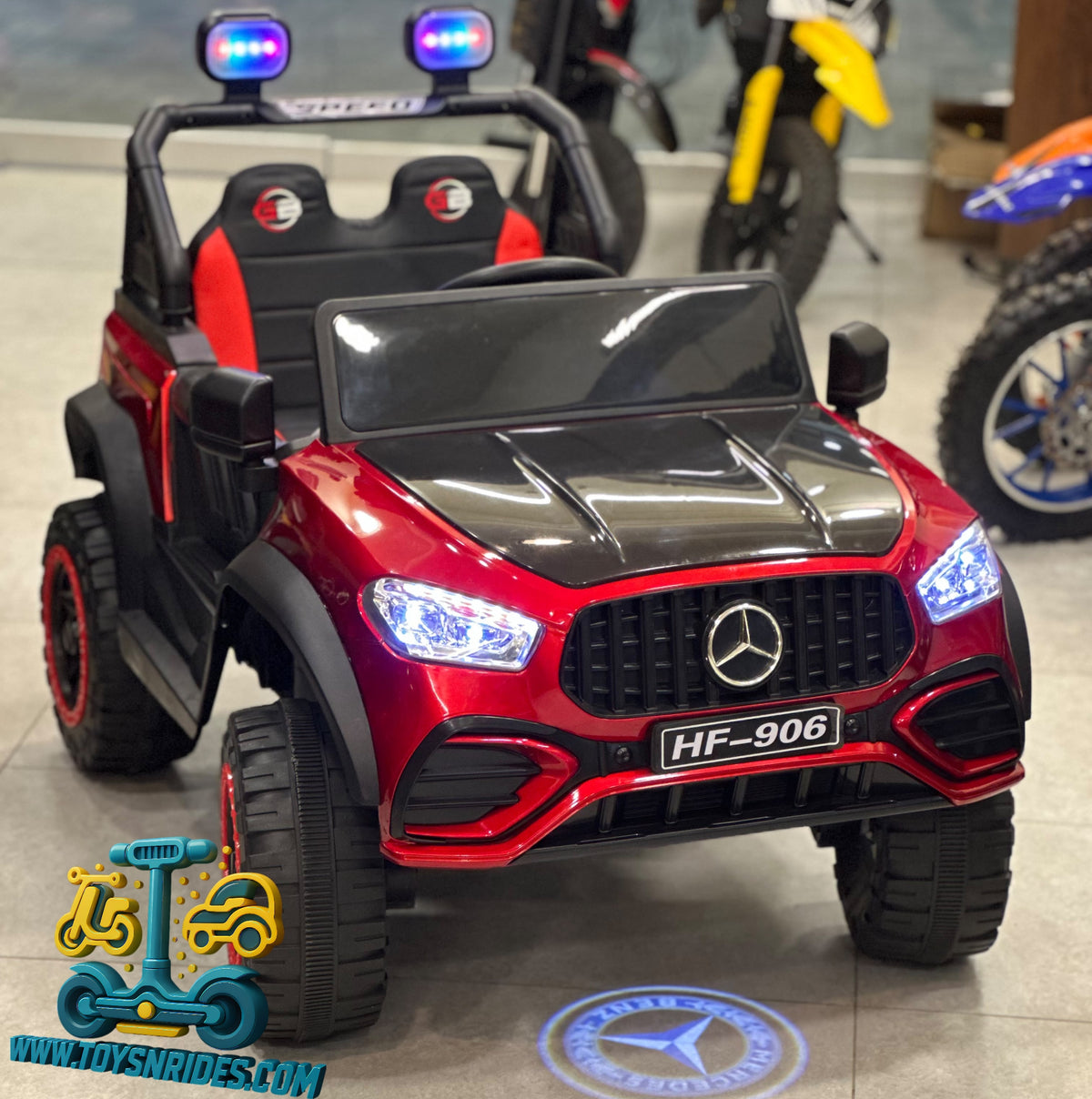 A red and black Mercedes-Benz style kids' ride-on truck with large off-road tires, blue LED headlights, and a license plate reading HF-906, photographed indoors in a showroom. A visible watermark/logo at bottom left reads www.