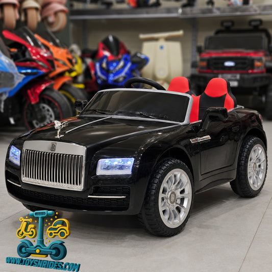 A black ride-on Rolls Royce toy car styled like a luxury Rolls-Royce, featuring a chrome grille, silver hood ornament, and red-and-black bucket seats, photographed in a showroom with other toy vehicles in the background.
